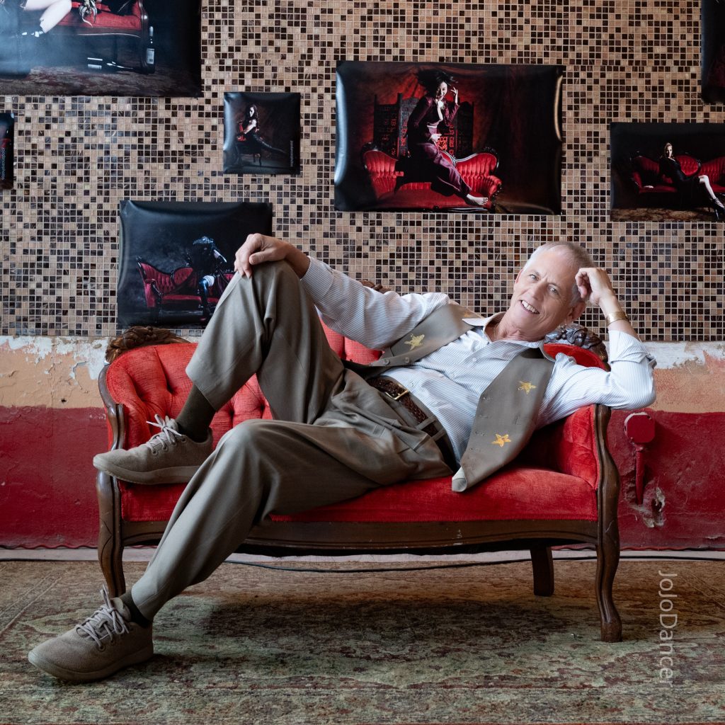 Rudi Harst reclining with guitar on a red sofa