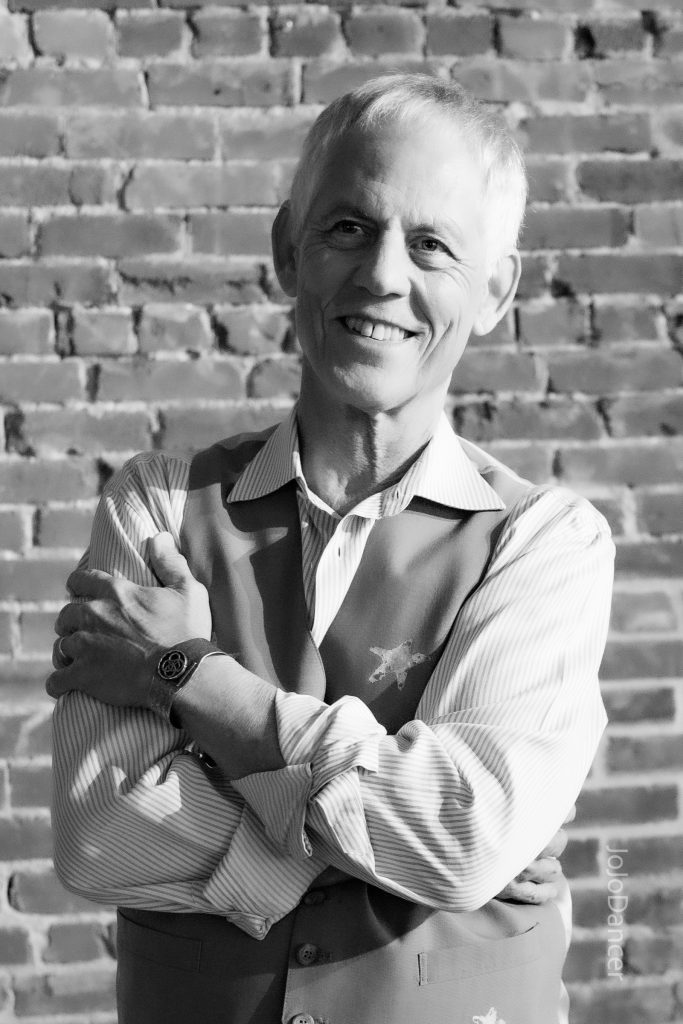 Rudi Harst smiling beside a brick wall