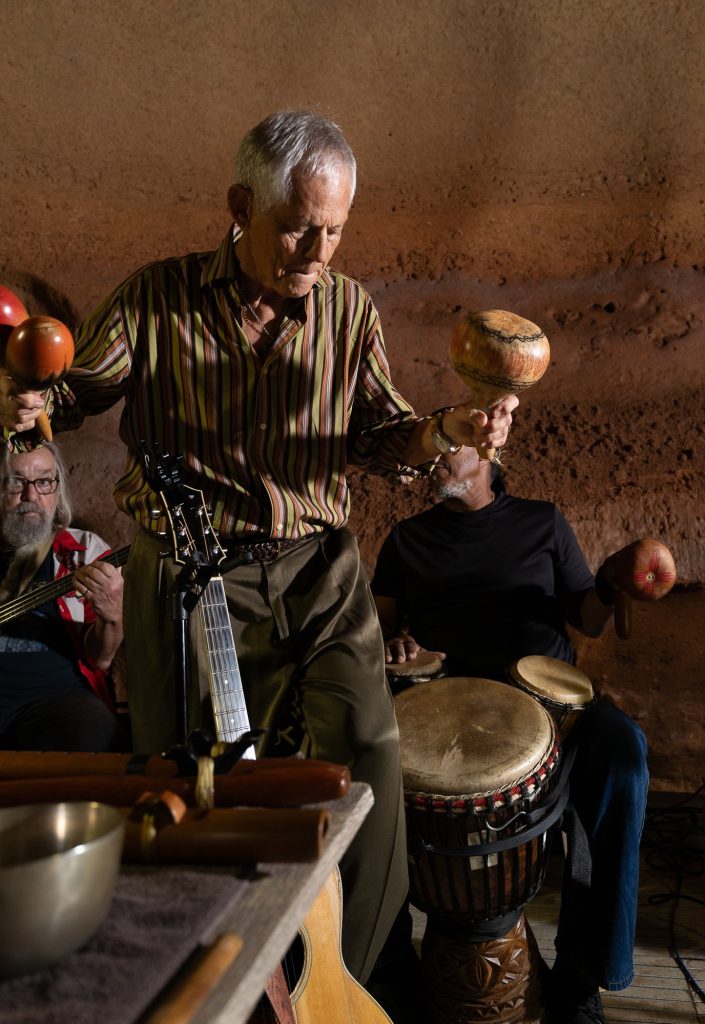 Rudi Harst holding two shakers beside hand drums against the cave wall