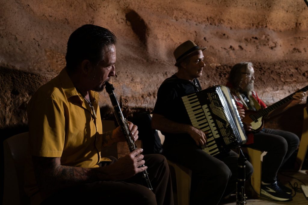 A wind player, accordionist and bassist seated against the warm cave wall