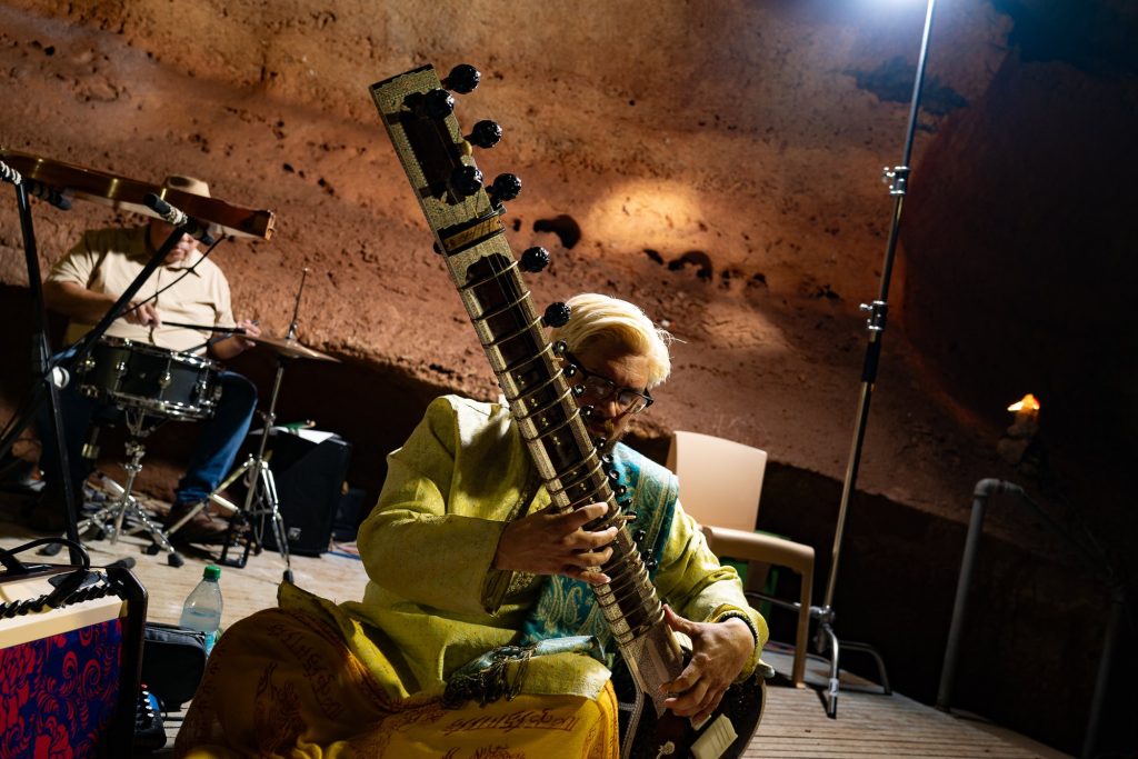 A seated sitar player in yellow clothing with a drummer behind him