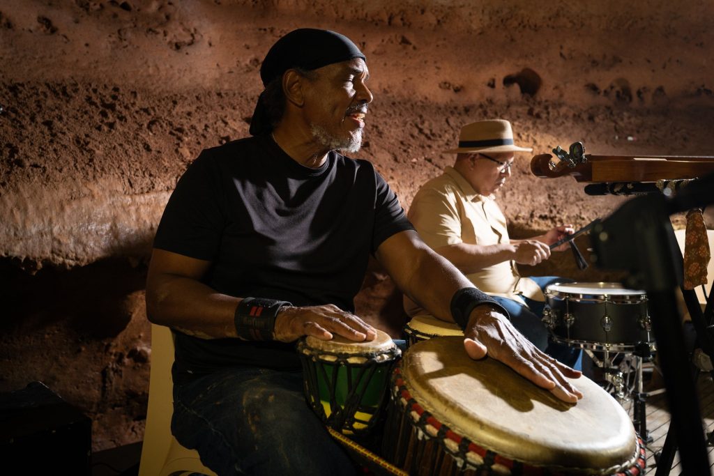 A smiling hand drummer beside a musician at a drum kit