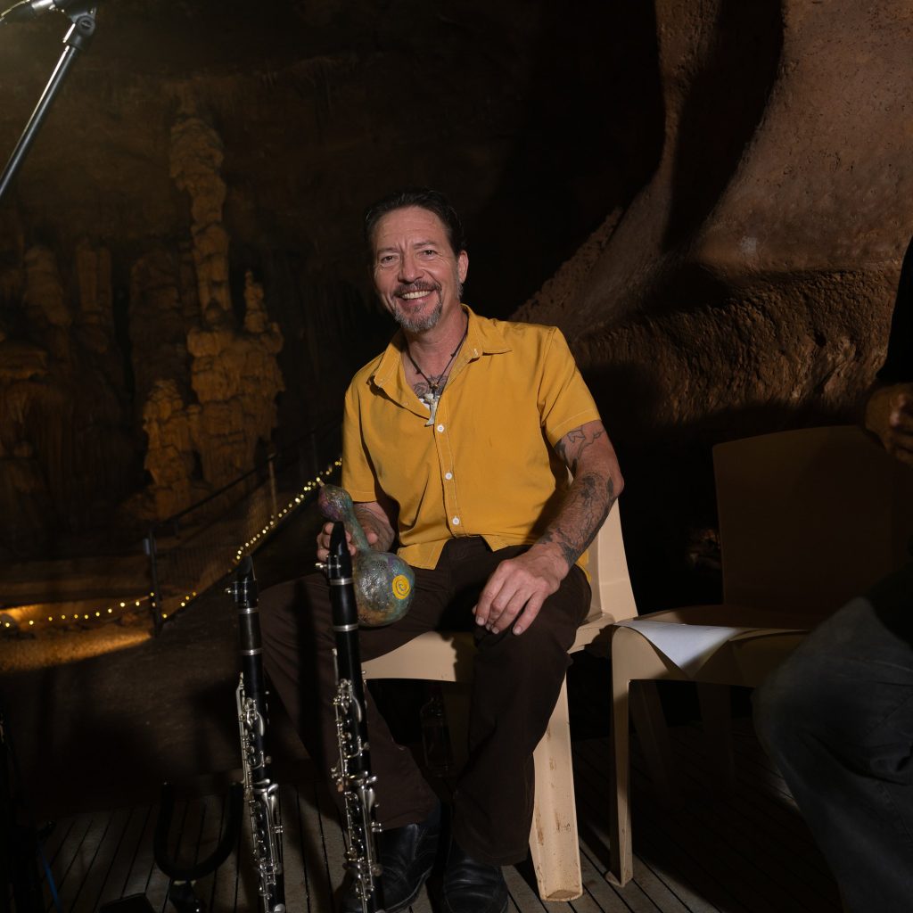 A smiling musician in a yellow shirt seated beside wind instruments