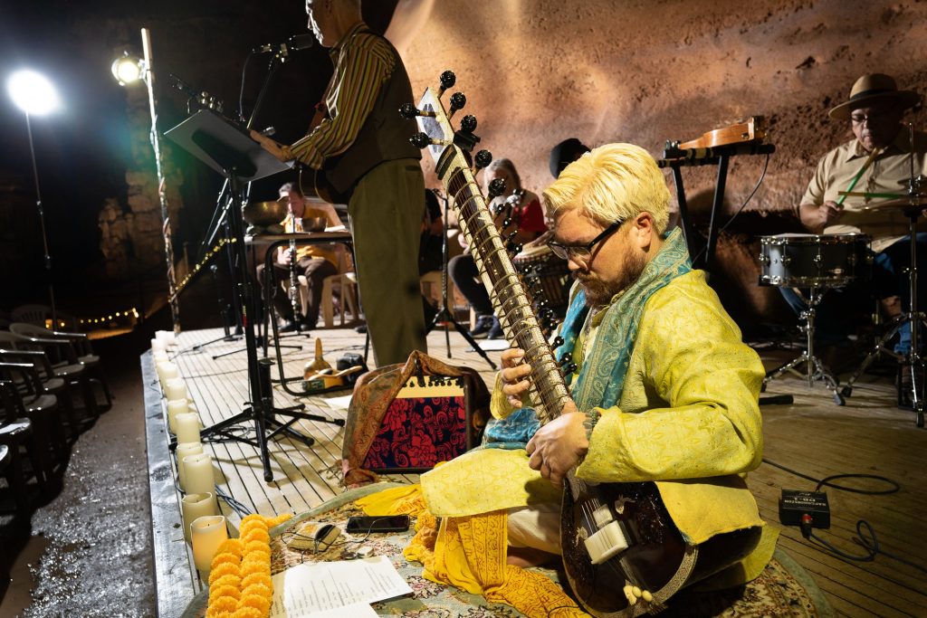 A seated sitar player performing beside a row of candles at the stage edge