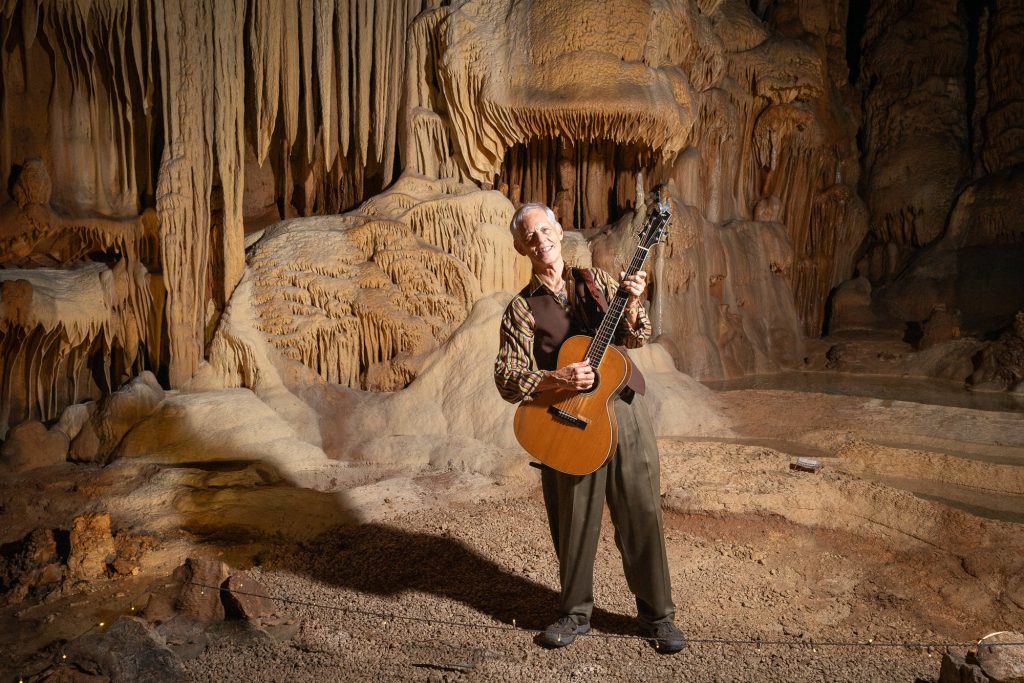 Wide portrait of Rudi Harst tilting his head with a guitar beneath cave formations