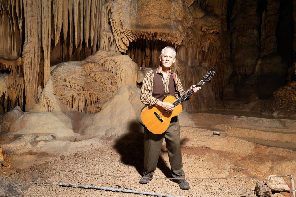 Rudi Harst standing with an acoustic guitar in front of layered cave formations
