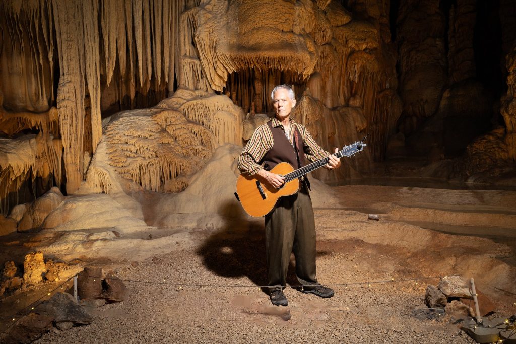 Full length portrait of Rudi Harst with guitar and a long shadow on the cave floor