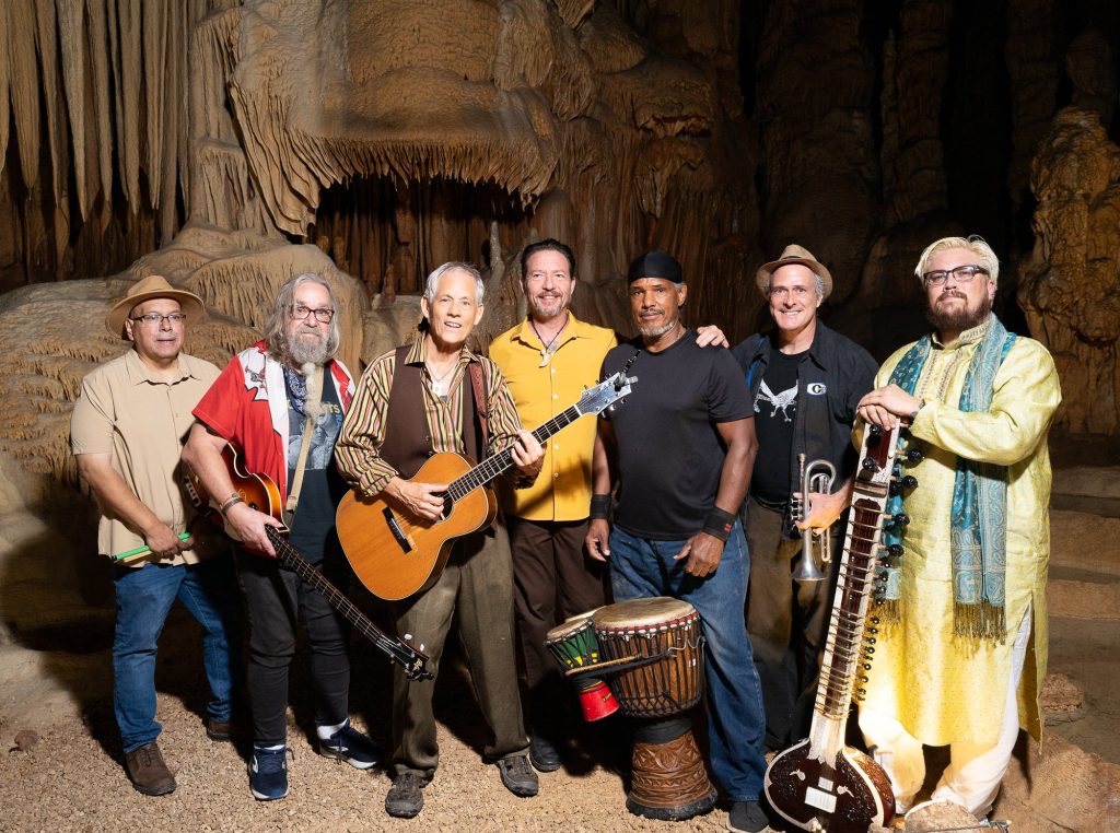 Wide group portrait of Rudi Harst and six fellow musicians beneath cave formations