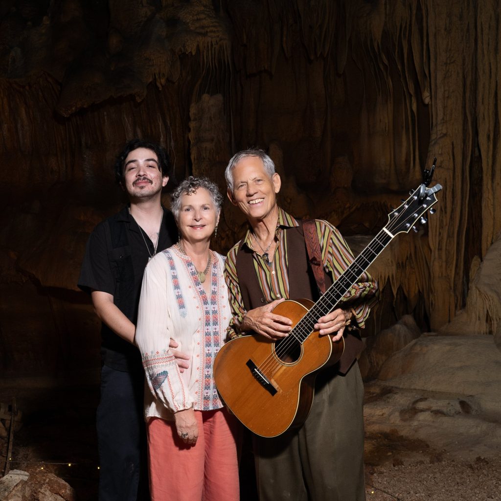 Rudi Harst holding his guitar and smiling beside two visitors