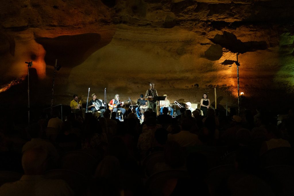 The ensemble performing across the cave stage beyond audience silhouettes