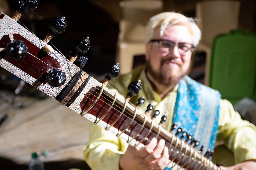 A sitar neck in sharp focus with its smiling player behind it