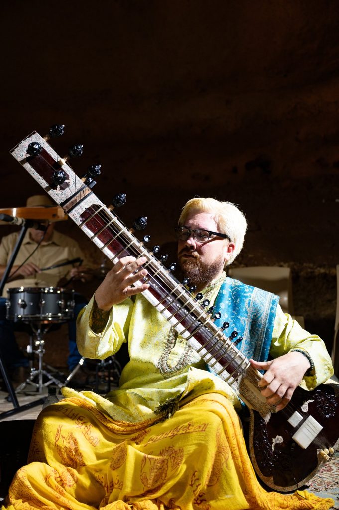A seated sitar player in yellow clothing looking toward the other musicians