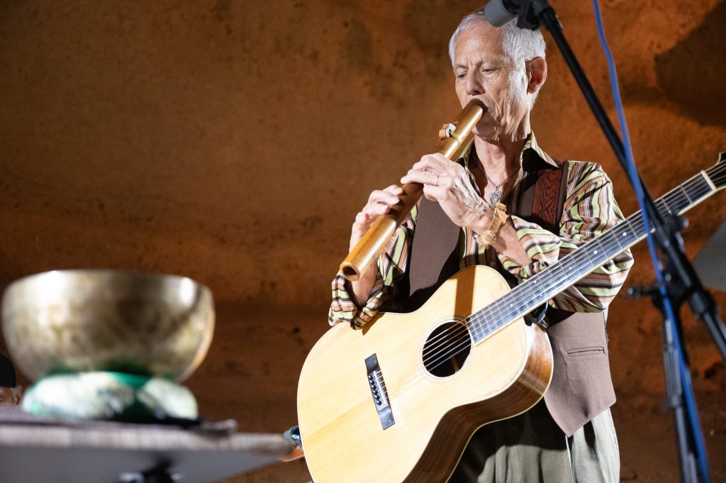 Rudi Harst playing a wooden flute with an acoustic guitar across his body
