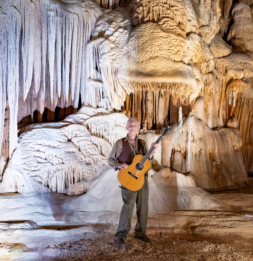 Vertical color portrait of Rudi Harst with guitar beneath illuminated cave formations