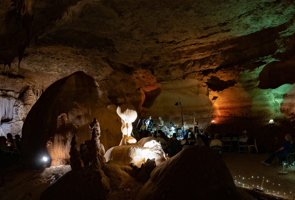 Wide cave interior with an illuminated formation and the concert stage beyond