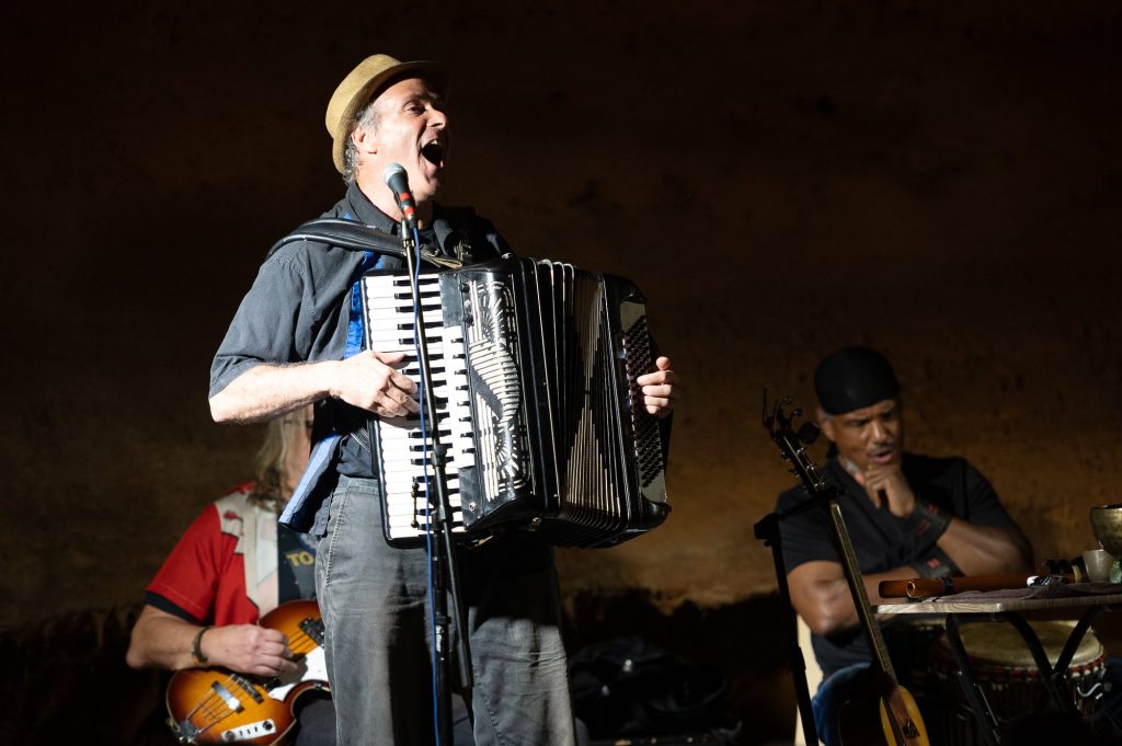 An accordionist singing into a microphone with the percussionist seated behind him